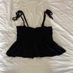 Svart linnetopp med knytband från Nelly - Svart linnetopp från Nelly med smockad överdel och volang nertill. Toppen har tunna axelband som knyts upptill för en söt och justerbar passform. Perfekt till varma dagar och enkel att styla med jeans eller kjol. Extra snygg om man stryker toppen innan användning😊