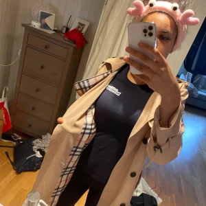 Beige trenchcoat med rutigt foder - Snygg beige trenchcoat med klassiskt rutigt foder på insidan. Jackan har bälte i midjan, knappar framtill och spännen vid ärmsluten. Perfekt för dig som vill ha en tidlös och stilren look. Fodret har svart, vitt och rött rutmönster som ger en lyxig känsla.