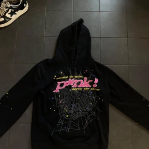 Spider hoodie  - Svart spider hoodie med stor huva och tryck på bröstet med texten 'Pink!' och spindelnätsmotiv. Små stjärnor i gult, rosa och blått över hela framsidan. På ryggen finns en gul stjärna. Perfekt för dig som gillar streetwear och unika prints.
