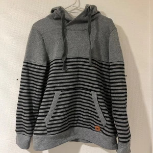 Grå och svart randig hoodie - Snygg grå hoodie med svarta horisontella ränder på nedre delen och ärmarna. Tröjan har huva med snörning och en stor magficka framtill. Mjuk och skön, perfekt för chill dagar. Insidan av huvan är också randig för extra stilpoäng.