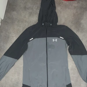 Svart och grå vindjacka från Under Armour - Snygg vindjacka från Under Armour i svart och grått med huva och hel dragkedja. Jackan har reflekterande detaljer och logga på bröstet samt stort Under Armour-tryck på ryggen. Perfekt för blåsiga dagar och sportiga outfits.