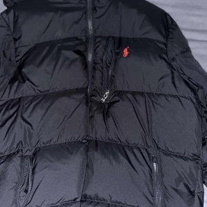 Svart pufferjacka från Polo Ralph Lauren - Säljer en svart pufferjacka från Polo Ralph Lauren med klassisk röd logga på bröstet. Jackan har dragkedja framtill, två sidofickor med dragkedja och quiltad design. Perfekt för kalla dagar och riktigt snygg till streetwear-stilen.