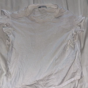 Vit volangärmad topp från Zara - Säljer en vit topp från Zara med korta ärmar och snygga volangdetaljer längs axlarna. Toppen har rund halsringning och är gjord i mjuk bomull, perfekt för en fräsch och enkel look. använd typ en gång utanför hemmet