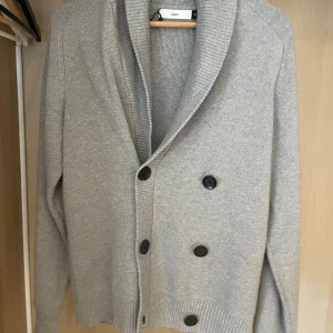 Grå kofta från Bläck med knappar - Snygg grå kofta från Bläck med dubbelknäppning och bred krage. Koftan är stickad i mjukt material och har långa ärmar samt ribbade muddar. Perfekt för lager-på-lager och chill dagar.