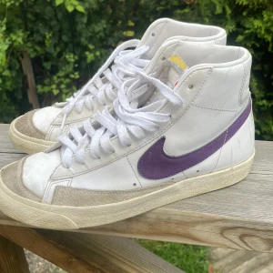 Nike Blazer Mid vit/lila sneakers - Nike Blazer Mid sneakers i vitt skinn med lila swoosh och lila Nike-logga på hälen. Klassisk retrodesign med högt skaft, vit snörning och beige mockadetaljer vid tån. Snygg kontrast mellan vitt och lila, perfekt för dig som gillar streetstyle. 