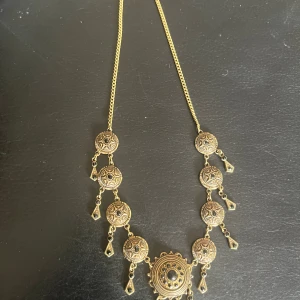 Guldigt statement-halsband med svarta stenar - Unikt halsband i guldton med flera runda medaljonger och svarta stenar. Kedjan är tunn och medaljongerna har detaljerade mönster och små hängande droppar med svarta stenar. Perfekt för dig som vill sticka ut med ett smycke som har vintagekänsla.