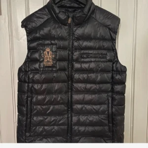 Svart dunväst från Morris - Säljer en äkta svart dunväst från Morris med quiltad design och hög krage. Västen har dragkedja framtill, två sidofickor och en bröstficka.