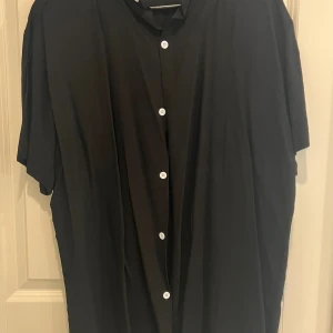 Svart kortärmad henley skjorta XL - Svart henley skjorta i storlek XL med korta ärmar och vita knappar framtill. Skjortan har en enkel och clean look med rak passform och mjukt tyg. Perfekt för dig som gillar minimalistisk stil och vill ha något som känns både chill och stilrent.
