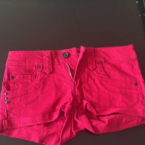 Röda jeansshorts från Terranova XXS - Snygga röda jeansshorts från Terranova i storlek XXS. Modellen är lågmidjad med klassiska fem fickor, nitar och bälteshällor. Materialet är kraftig bomull med dragkedja och knapp framtill. Perfekta för varma dagar och en färgstark look.