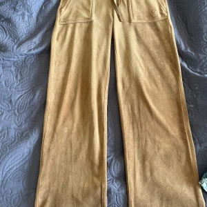 Juicy Couture mjukisbyxor XS brun - Supermysiga bruna mjukisbyxor från Juicy Couture med broderad logga, stora fickor framtill och snörning i midjan med silver detaljer. Byxorna har en rak passform och är gjorda i mjuk velour som känns lyxig och skön mot huden.