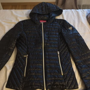 Svart quiltad dunjacka från Guess - Svart dunjacka från Guess med quiltad design och silverfärgade dragkedjor. Jackan har huva, två fickor med dragkedja och en smickrande passform. Perfekt för kyliga dagar när du vill vara både varm och snygg. Sitter tajt vid midjan 