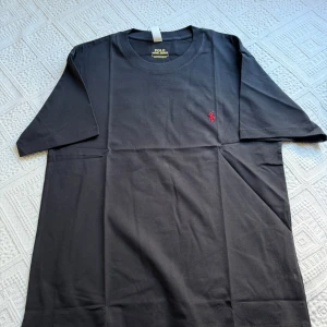 Svart t-shirt från Polo Ralph Lauren - Svart t-shirt från Polo Ralph Lauren med klassisk röd broderad logga på bröstet. T-shirten har rund halsringning och är tillverkad i mjuk bomull. Passformen är slim och modellen är kortärmad, perfekt för en clean och stilren look.