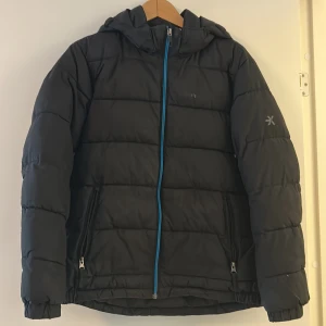 Svart pufferjacka från Everest 164 - Svart pufferjacka från Everest med huva och blå dragkedja. Jackan har två fickor med dragkedja och diskret logga på ärmen. Perfekt för kalla dagar och har en sportig vibe. Snyggt quiltad och med elastiska muddar vid ärmslut. Jackan är i bra skick!