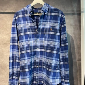 Ralph Lauren skjorta - Snygg blårutig skjorta från Ralph Lauren med klassisk button-down krage och broderad logga på bröstet. Skjortan har långa ärmar, vita knappar och är tillverkad i mjuk bomull. Perfekt för dig som gillar en stilren och tidlös look.