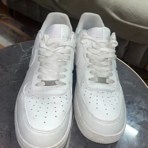 Nike Air Force 1 helvita sneakers - Klassiska Nike Air Force 1 sneakers i helvitt skinn med vit sula och snörning. Ikonisk siluett med perforerad tå och Nike-logga på sidan. Perfekta för dig som vill ha en clean och tidlös look. Passar till allt och är ett måste i garderoben.