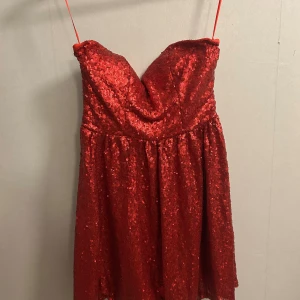 Röd paljettklänning Model Behaviour - Glittrig röd kortklänning från Model Behaviour med paljetter över hela klänningen. Axelbandslös modell med hjärtformad urringning och dragkedja bak. Perfekt för dig som vill sticka ut och glänsa. Fodrad insida för extra komfort.