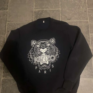 Svart sweatshirt från Kenzo med tiger - Svart sweatshirt från Kenzo med ikoniskt broderat tigermotiv och texten 'Kenzo Paris' framtill. Tröjan har rund halsringning och långa ärmar. Perfekt statement-plagg för dig som gillar streetstyle och snygga detaljer.