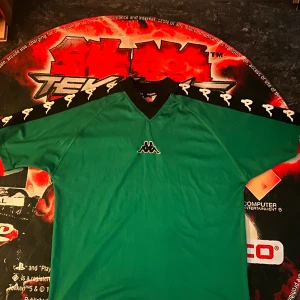 Grön Kappa fotbollströja retro - Retro grön fotbollströja från Kappa med svart V-ringad krage och klassiska Kappa-loggor längs axlarna. Tillverkad i lätt och ventilerande material, perfekt för träning eller match. Snygg och sportig design med tydlig logga på bröstet.