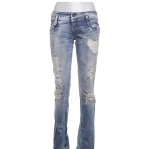 Ljusblå slitna jeans med bootcut - Säljer ett par ljusblå jeans med slitningar och trasiga detaljer på benen. Jeansen har klassisk femficksdesign, låg midja och bootcut-ben. Perfekta för en avslappnad och trendig look.