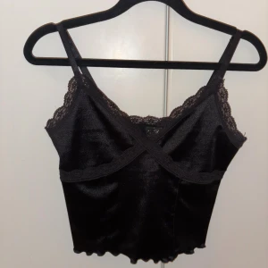 Svart satinlinnetopp med spets H&M - Supersnygg svart linnetopp från H&M Divided i satinliknande material med spetsdetaljer längs urringning och axelband. Croppad modell med vågig kant nertill och smala justerbara axelband. Perfekt att styla med jeans eller kjol för en trendig look.