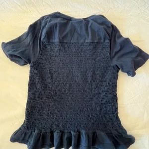 Mörkblå blus med volang från Gina Tricot - Supersöt mörkblå blus från Gina Tricot med korta ärmar, smockad baksida och volangkant nedtill. Perfekt för dig som gillar en feminin och trendig stil. Tillverkad i mjuk bomull som känns skön mot huden.