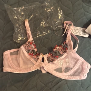 Rosa blommig spets-bh från Freya - Supersöt bh i ljusrosa mesh från Freya med broderade blommor i rött och svart på kuporna. Justerbara axelband med gulddetaljer och liten rosett framtill. Perfekt för dig som gillar romantiska detaljer och blommigt mönster.