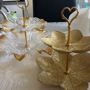 Två fina fat som man kan servera fika och annat gott på, bra skick använt få gånger - Bilderna visar två eleganta våningsfat, ett i glas med gulddetaljer och ett i metall med bladformad design och guldfinish. Perfekta för servering av kakor eller frukt vid festliga tillfällen. Båda ser ut att vara i mycket gott skick utan synliga defekter eller slitage.