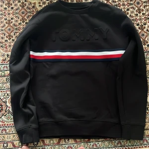 Svart Tommy Hilfiger sweatshirt 164 - Svart sweatshirt från Tommy Hilfiger med upphöjd TOMMY-text på bröstet och en bred rand i marinblått, vitt och rött över bröstet. Klassisk rund halsringning och långa ärmar. Perfekt för dig som gillar stilrena och sportiga plagg.