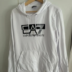 Vit hoodie från Emporio Armani EA7 - Snygg vit hoodie från Emporio Armani EA7 med svart logotryck på bröstet. Tröjan har huva med snörning, stor magficka och långa ärmar.