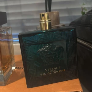 Versace Eros Eau de Toilette - Versace Eros Eau de Toilette med en stilren, fyrkantig flaska i mörk turkos glas och guldigt sprayhuvud. Flaskan har ett klassiskt Medusa-huvud i relief och geometriskt mönster. En ikonisk doft som sticker ut i badrumshyllan.
