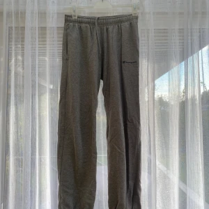 Grå mjukisbyxor från Champion - Gråa mjukisbyxor från Champion med elastisk midja och sidofickor. Byxorna har en avslappnad passform och är perfekta för chill dagar. Liten Champion-logga broderad på ena benet. Tillverkade i mjukt bomullsmaterial.