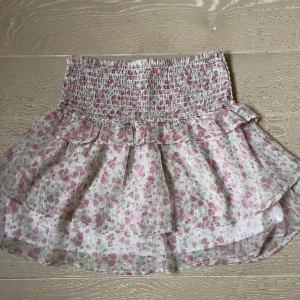 Blommig volangkjol - Supersöt kort kjol med blommigt mönster i rosa och grönt på vit botten. Kjolen har smock i midjan och volangdetaljer. 