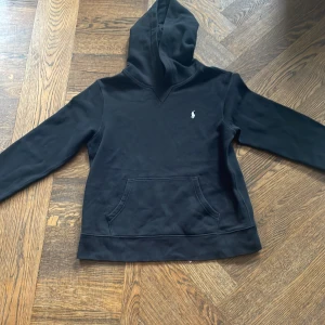 Svart hoodie från Polo Ralph Lauren - Svart hoodie från Polo Ralph Lauren med klassisk vit broderad logga på bröstet. Tröjan har huva, känguruficka och långa ärmar. Mjuk insida i fleece och stilren design som funkar till jeans eller joggers. Perfekt för chill eller häng med kompisar.