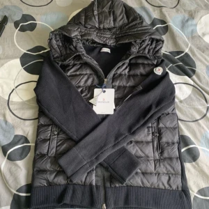 Svart Moncler pufferjacka med huva - Säljer en svart pufferjacka från Moncler med stickade ärmar och quiltad kropp. Jackan har huva, dragkedja framtill och två fickor med knappar. Perfekt för dig som vill ha en snygg och sportig look med premiumkänsla.