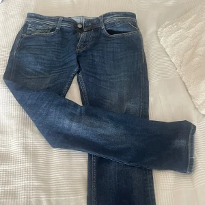 Blå Replay Rocco jeans straight fit - Säljer ett par snygga Replay Rocco jeans i klassisk blå tvätt med slitningar och raka ben. Jeansen har fem fickor, tydliga kontrastsömmar och läderpatch bak i midjan. Köpta för 1800kr och använda ett fåtal gånger. 