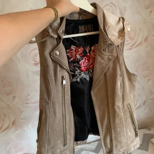 Beige mockaväst med huva från Jofama - Snygg beige mockaväst från Jofama by Caroline Roxy med avtagbar huva, dragkedjor framtill och coola nitar på ryggen. Insidan har broderade rosor i rött och rosa. Västen har detaljer som spännen i sidorna och en bikerinspirerad look.