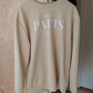 Beige sweatshirt med Paris-tryck - Snygg beige sweatshirt från Sundays med stort vitt Paris-tryck på bröstet och texten 'Monet's garden at Giverny, Ville Romantique'. Tröjan har rund hals, långa ärmar och ribbade muddar. Perfekt för dig som gillar chill och trendiga prints.