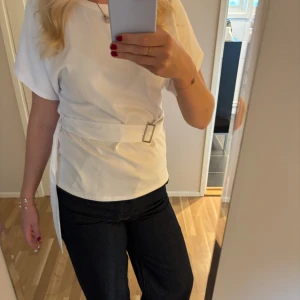 Vit topp med bälte från Zara - Säljer en stilren vit topp från Zara med korta ärmar och rund hals. Toppen har ett brett bälte i midjan med silverfärgat spänne som ger en snygg siluett. Perfekt att matcha med jeans eller kjol för en clean look. 💞💞☺️