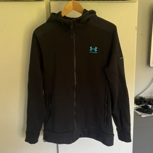 Svart hoodie från Under Armour M - Svart hoodie från Under Armour med blå logga på bröstet och ärmen. Tröjan har dragkedja hela vägen, två fickor och huva. Perfekt för träning eller chill. Materialet är mjukt och känns sportigt, med långärmad passform och normal passning.