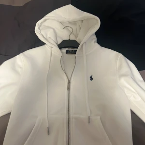 Vit zip hoodie från Polo Ralph Lauren - Säljer en stilren vit hoodie från Polo Ralph Lauren med dragkedja och huva. Tröjan har snörning vid huvan, två fickor framtill och klassisk blå broderad logga på bröstet. Perfekt för en clean och avslappnad look. Storlek S, nypris 1899kr, använd endast 2 gånger, priset kan diskuteras.