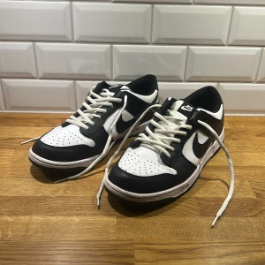 Nike Dunk Low svart/vit skinn - Nike Dunk Low sneakers i svart och vitt skinn med klassisk swoosh på sidorna. Skorna har snörning, perforerad tåbox och platt sula. Perfekt för dig som gillar streetstyle och vill ha en ikonisk sneaker i garderoben.