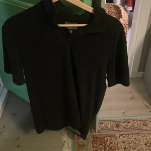Svart pikétröja från H&M, M - Klassisk svart pikétröja från H&M i regular fit. Tröjan har korta ärmar och krage, perfekt för en clean och enkel stil. Materialet känns mjukt och bekvämt mot huden. Passar dig som gillar en stilren look med avslappnad känsla.