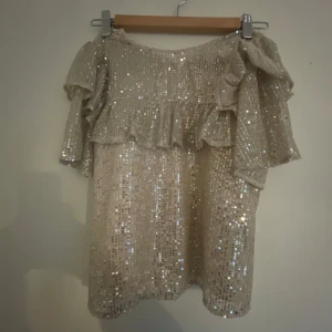 Super fin Glittrig volangblus från Lindex - Supersnygg glittrig blus från Lindex i ljusbeige med paljetter över hela toppen. Blusen har korta volangärmar och en lös passform som ger en snygg och festlig vibe. Perfekt för dig som vill sticka ut med extra glitter och volanger!(helt perfekt till fester, nyår och massa andra tillfällen)