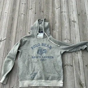 Grå hoodie Polo Bear Ralph Lauren - Grå hoodie från Polo Ralph Lauren med Polo Bear-tryck och texten 'Ralph Lauren 1967' i blått framtill. Tröjan har huva med snörning, känguruficka och mjukt fleecetyg på insidan. Perfekt för en chill och avslappnad stil.