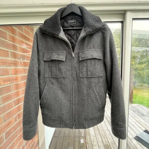 Grå ulljacka från Eastwest, S - Snygg grå jacka från Eastwest i ullblandning med ribbad krage och quiltat foder. Jackan har dragkedja framtill, fyra fickor med lock på bröstet och snedställda fickor vid sidorna. Perfekt för kyliga dagar och ger en clean, stilren look.