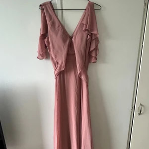 Ljusrosa långklänning med volang - Säljer en ljusrosa långklänning med v-ringad front och volangdetaljer vid axlarna. Klänningen har korta ärmar, omlottkänsla och ett knytband i midjan samt dragkedja i bak. 