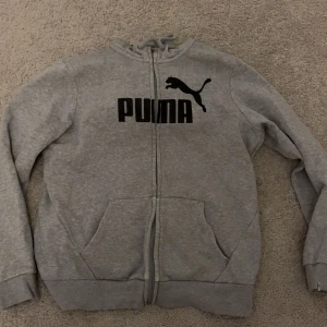 Grå hoodie från Puma med dragkedja - Säljer en grå hoodie från Puma med svart logga på bröstet. Tröjan har huva med snörning, hel dragkedja och två stora fickor framtill. Perfekt för chill dagar eller när du vill ha en sportig look.