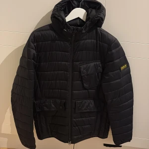 Svart Barbour pufferjacka med huva - Svart pufferjacka från Barbour International med huva och quiltad design. Jackan har praktiska fickor med knappar, dragkedja framtill och en snygg Barbour-patch på ärmen. Tillverkad i polyamid med polyesterfyllning, perfekt för kalla dagar.