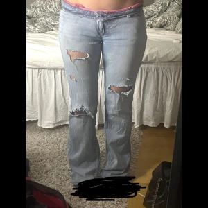 Ljusblå slitna bootcut jeans - Säljer ett par ljusblå låg midjade jeans med bootcut passform och snygga slitningar på båda benen. Jeansen har klassisk femficksdesign och låg midja. Perfekta för dig som gillar en avslappnad och trendig look. 