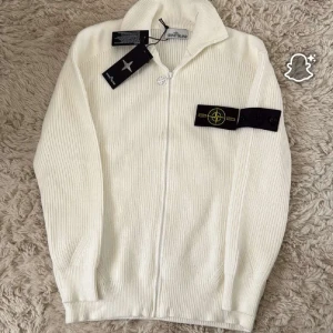 Vit stickad kofta från Stone Island - Vit stickad kofta från Stone Island med hel dragkedja och klassisk logga på ärmen. Tröjan har ribbad struktur, hög krage och snygga detaljer som gör den riktigt clean. Perfekt för dig som gillar stilrena och exklusiva plagg.